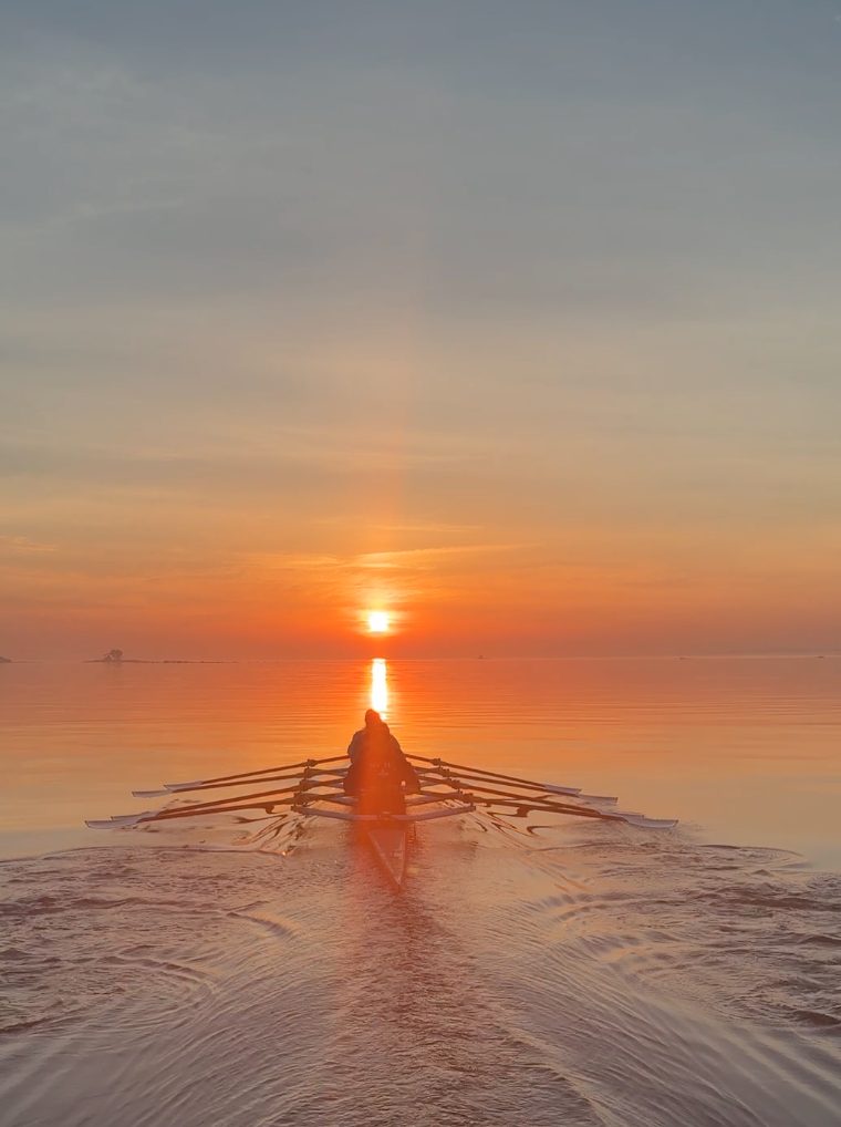 A sculler rowing straight into a rising orange sun on dead-calm water