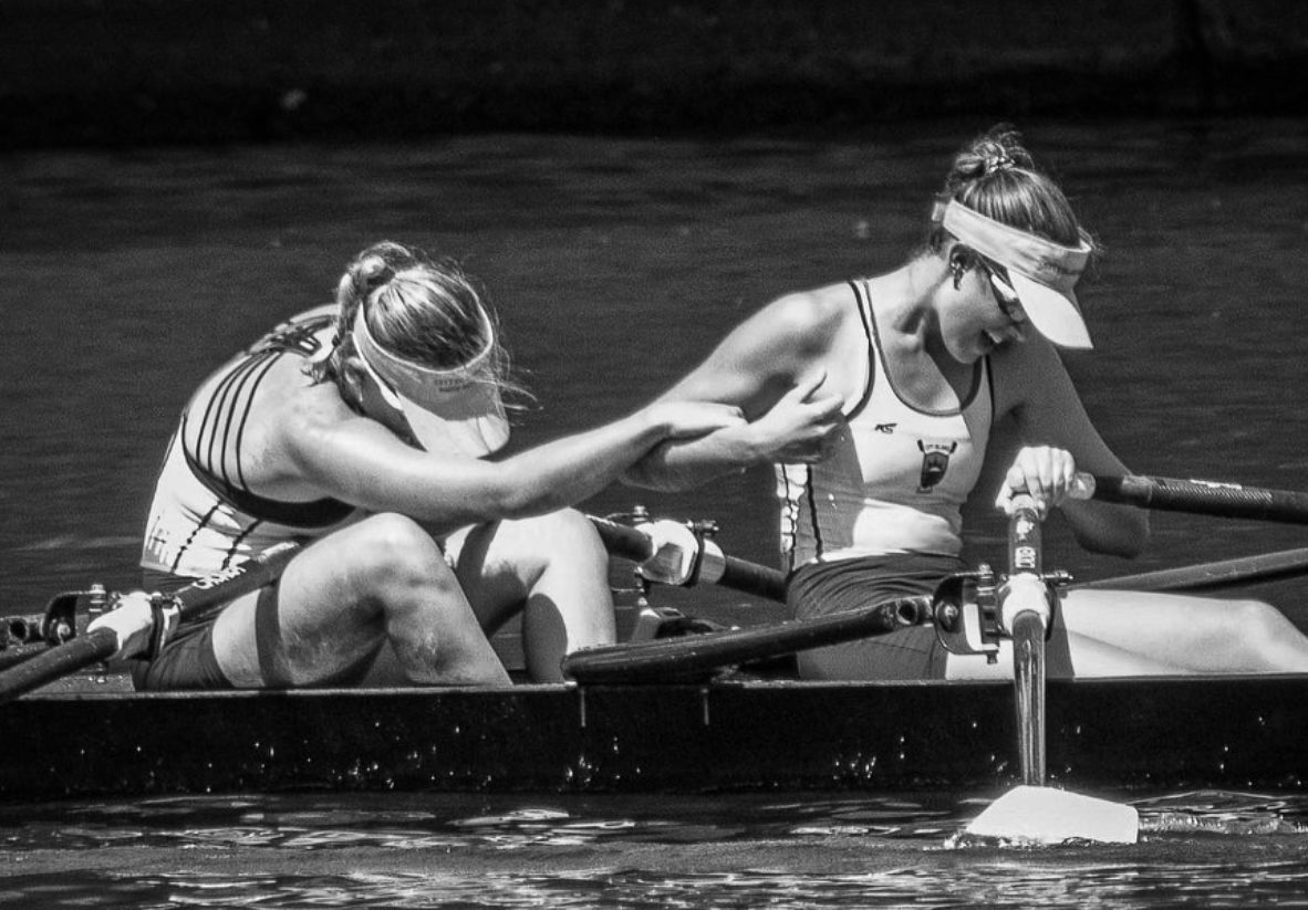 Two racers in the boat, one reaching over to her partner, in black and white